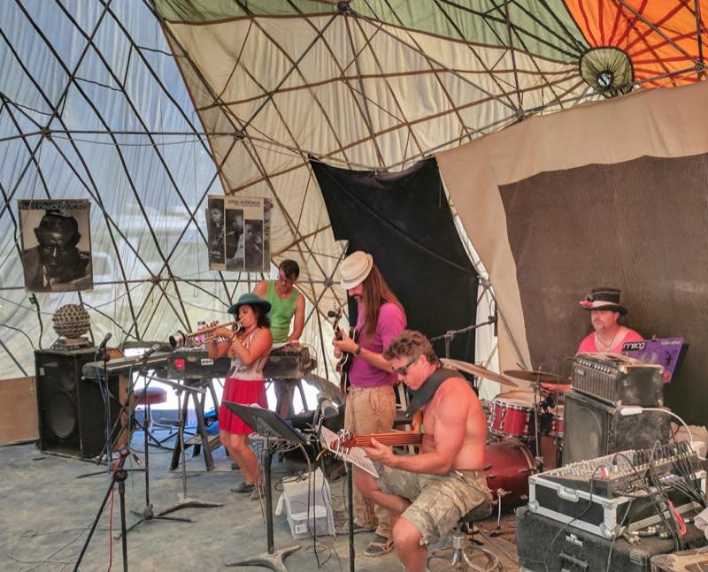 A band mid set under a geodesic dome, trumpet and keys and guitar and drums, with Thelonious Monk and John Coltrane posters pinned to the canvas behind them.
