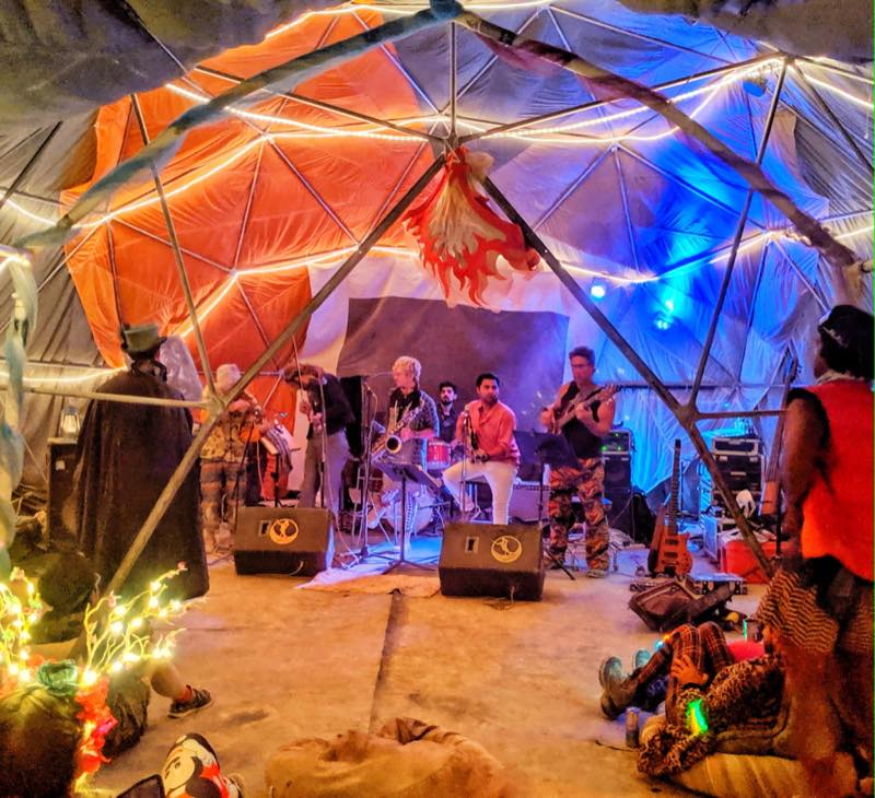 A night set inside the dome, its struts traced in orange and blue light, a saxophonist mid phrase while the audience sits on the ground.