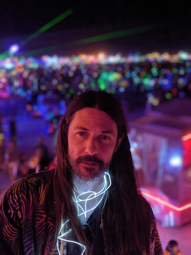 A portrait at night, electroluminescent wire glowing at the collar, green laser lines overhead and a blurred field of coloured lights behind.