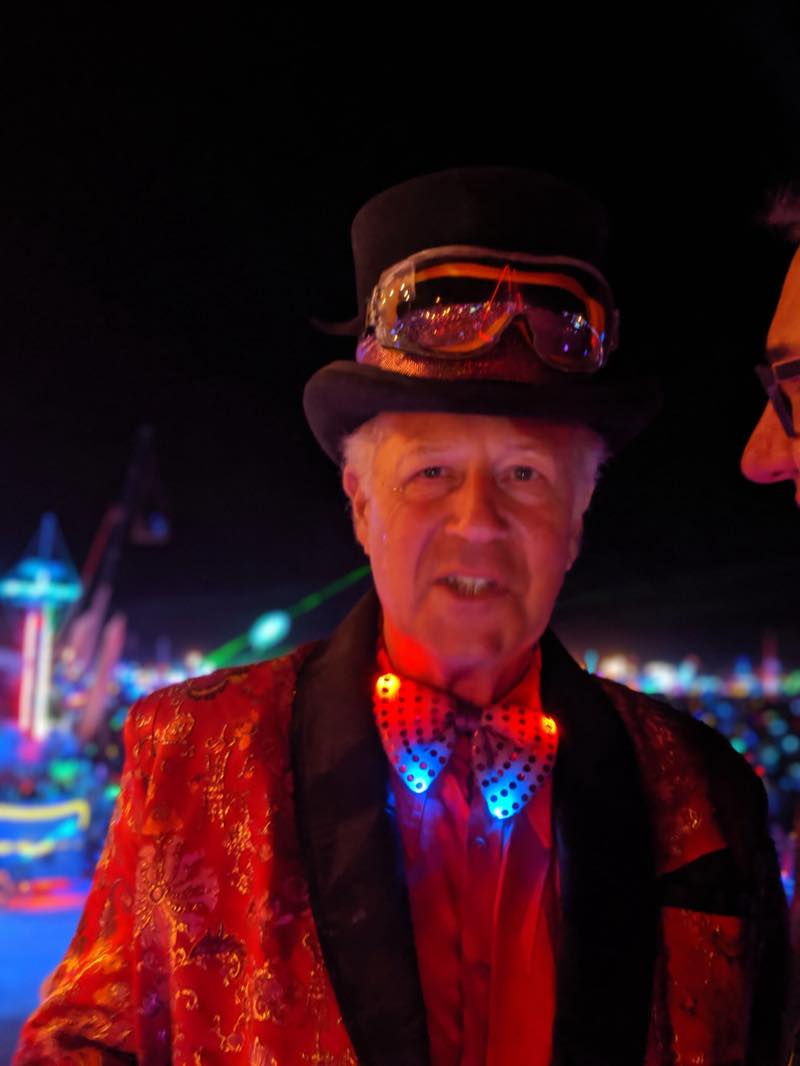 Cabro at night in a black top hat with dust goggles pushed up on the brim, a red brocade jacket, and a bow tie strung with tiny lights.