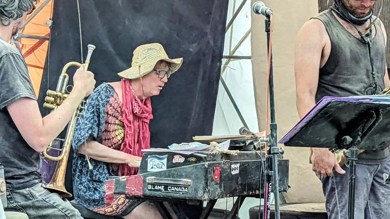 A keyboardist in a straw hat and red scarf plays a keyboard mounted on a battered road case stencilled BLAME CANADA, a trumpeter alongside.