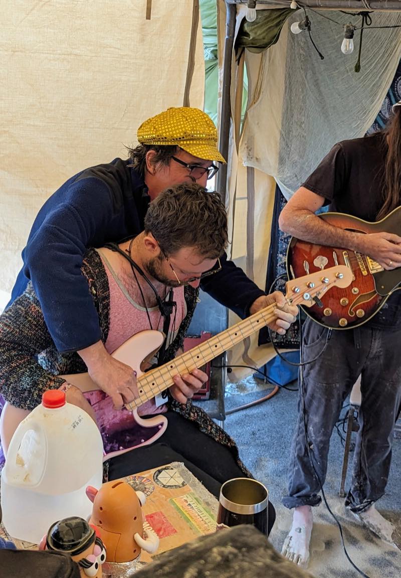 One player leans over another&rsquo;s shoulder to guide their hands on the neck of a pink bass, while a barefoot guitarist plays alongside in the dust.