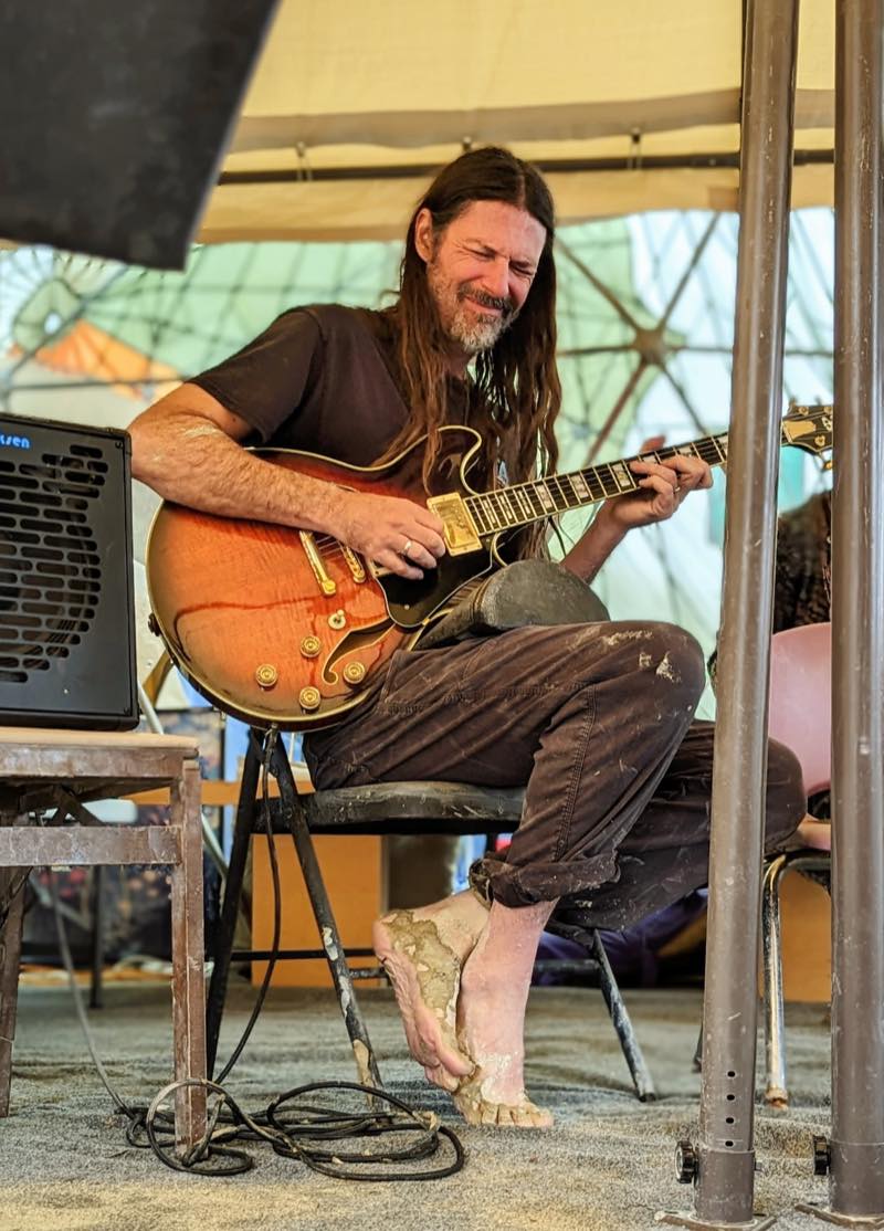 A guitarist with eyes closed mid phrase on a sunburst hollowbody, barefoot, both feet caked in dried playa mud.
