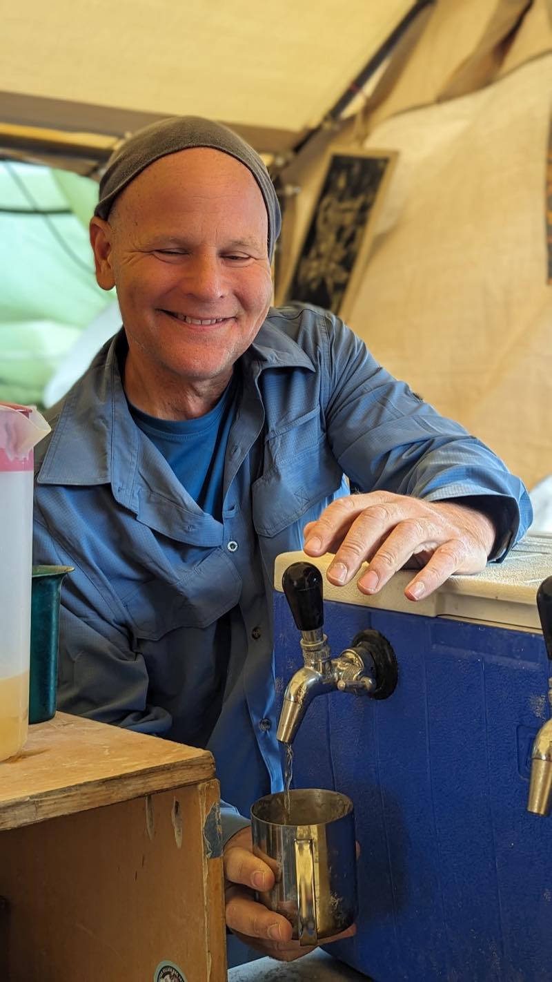 Sparky, grinning, draws a drink from the tap of a blue cooler into a dented metal mug.