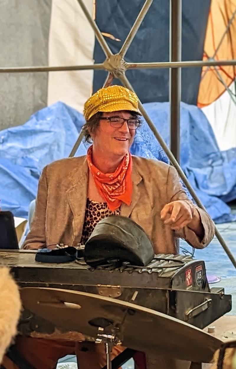 A keyboardist in a gold sequinned cap and orange bandana grins and points across the tent from behind the keys.