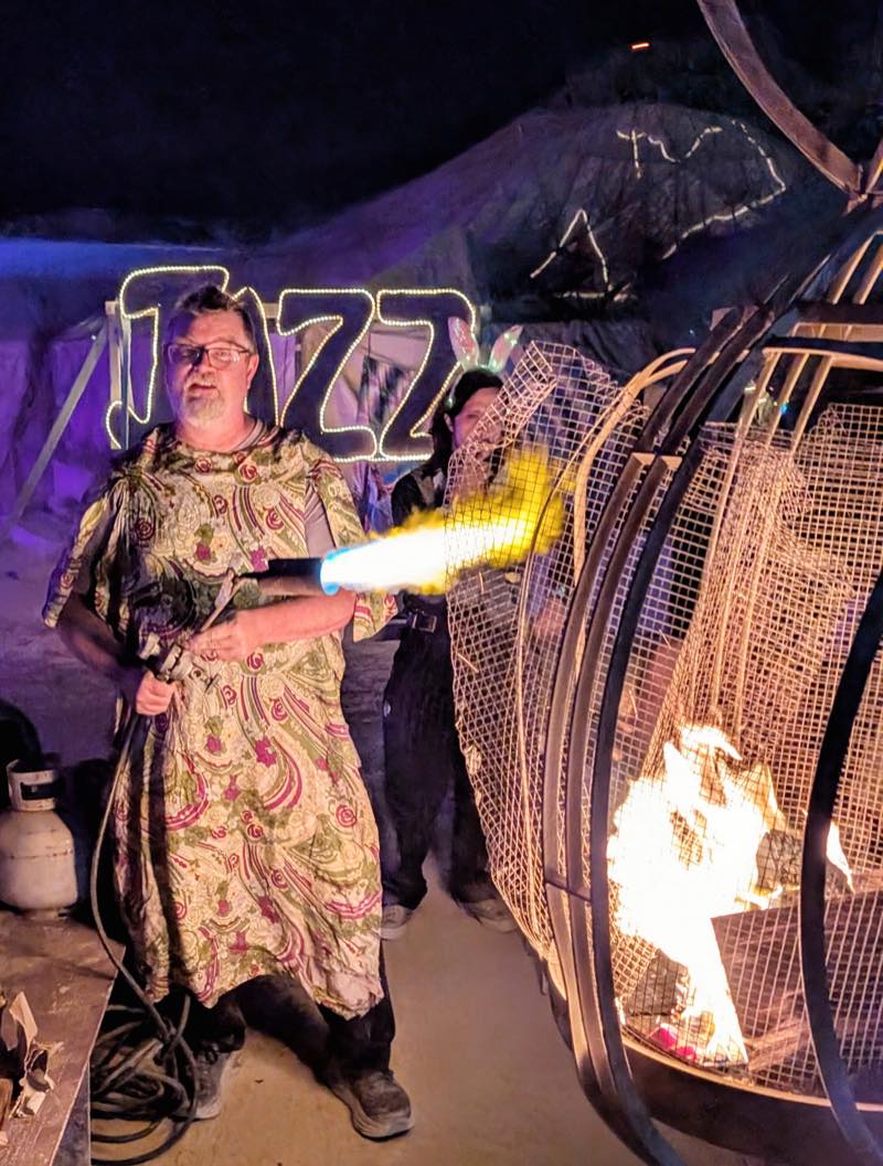 Someone in a floral dress fires a propane torch into a caged fire sculpture at night, the bulb lit word JAZZ glowing behind them.