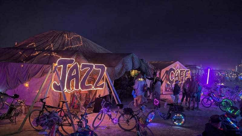 The camp under a violet night sky, JAZZ and CAFE in warm bulb lettering, bicycles with illuminated wheels parked across the sand.