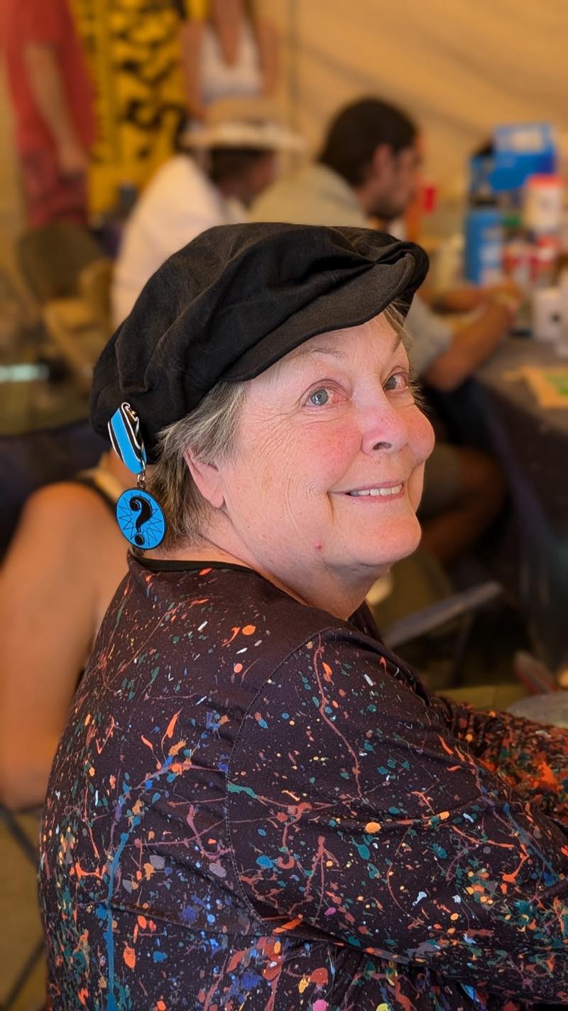 A portrait in the tent: a black newsboy cap, a bright blue question mark earring, and a paint splattered top, smiling back over one shoulder.