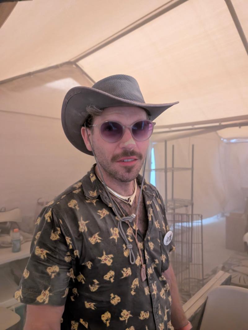 A portrait during build week: a grey outback hat, round purple sunglasses, and a Pikachu print shirt already thick with dust.