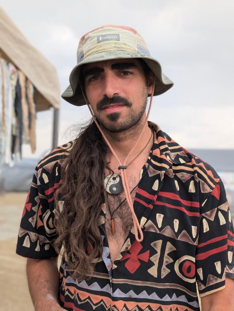 A portrait at the camp: an olive bucket hat, long hair, and a Playa Jazz Caf&eacute; yin yang medallion dated 2023 and stamped 6 & E.