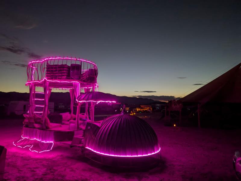 The camp structure at dusk, a two tier deck and a ribbed dome outlined in pink light, the mountains still visible behind.