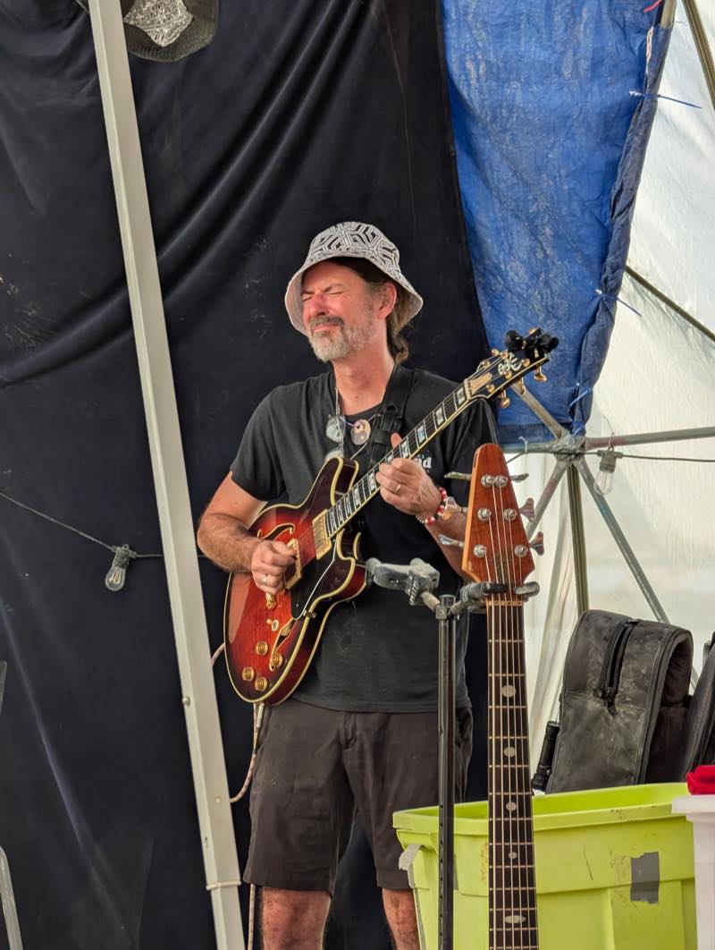 A guitarist in a patterned bucket hat plays a cherry red hollowbody with eyes closed, a bass waiting on a stand alongside.