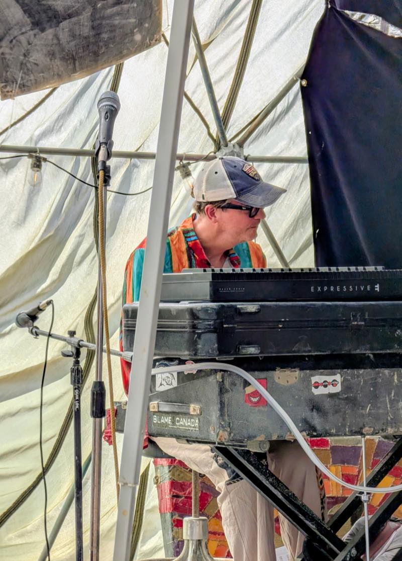 A keyboardist in a ball cap and sunglasses at the keys, the same battered road case still doing duty as a stand.