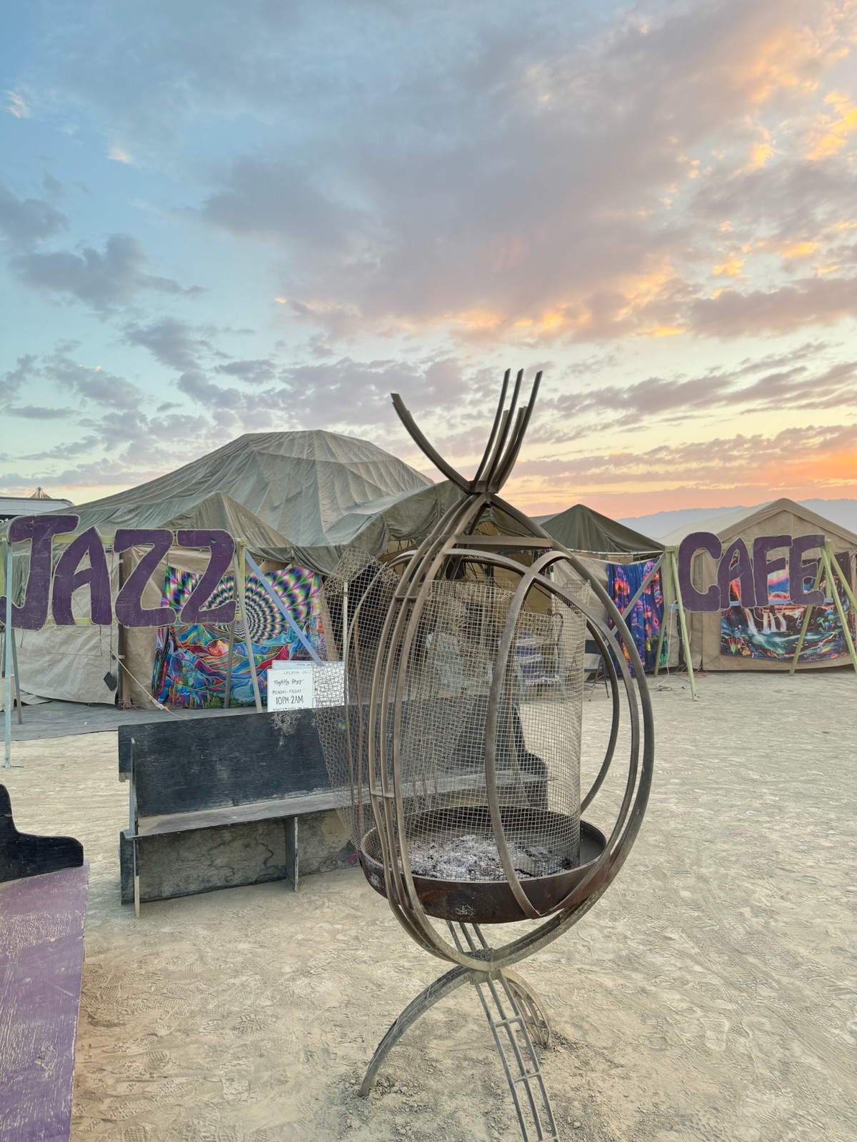 The camp at sunrise: hand-painted JAZZ CAFE letters, a fire sculpture, and the open playa beyond.